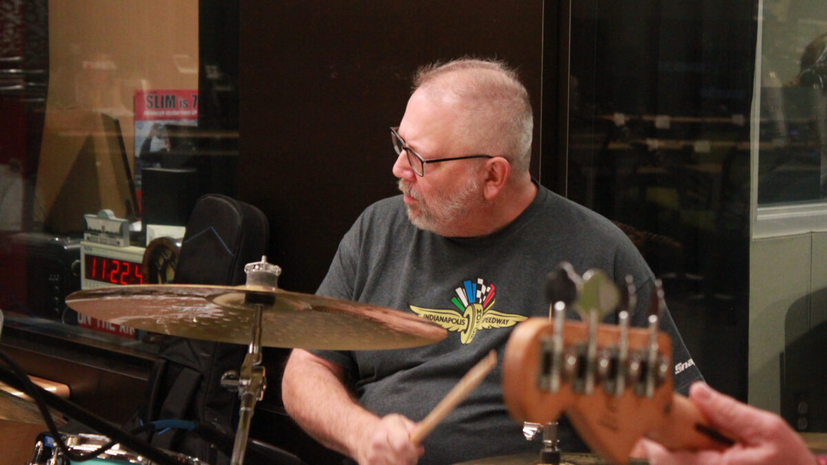 Slim-Forsythe-and-the-Boss-Stingrays-5 An adult playing a drum set in front of a radio studio