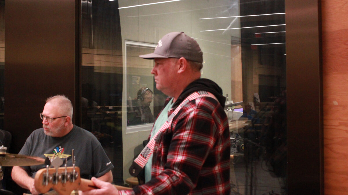 Slim-Forsythe-and-the-Boss-Stingrays-6 An adult playing a drum set and an adult playing electric bass in front of a radio studio