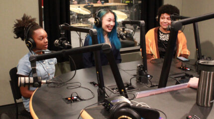 Two teens and an adult wearing headphones speaking into microphones in a radio studio
