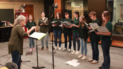 An adult conducting seven teens and an adult singing in a choir while holding binders