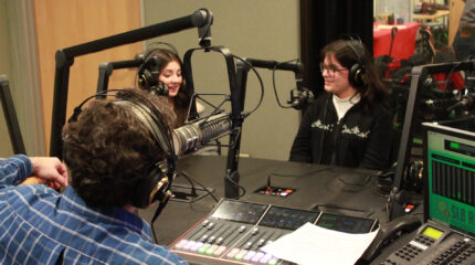 Two teens wearing headphones speaking into microphones being interviewed by an adult wearing headphones sitting behind a microphone and a mixer in a radio studio