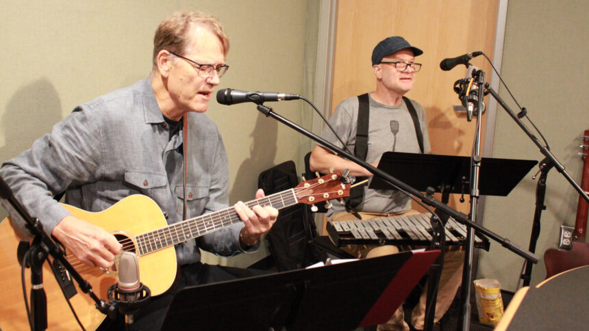 An adult playing acoustic guitar and singing into a microphone and an adult playing glockenspiel in a radio studio