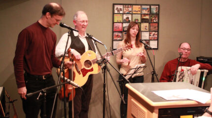 An adult holding a violin standing behind a microphone, an adult holding an acoustic guitar and speaking into a microphone, an adult speaking into a microphone and standing behind sheet music on a music stand, and an adult holding an accordion sitting behind a microphone in a radio studio