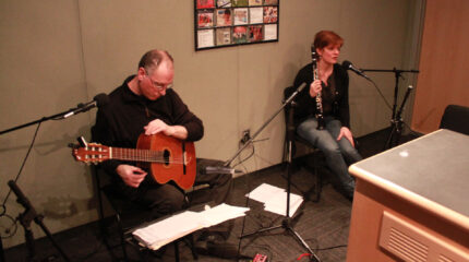 An adult holding an acoustic guitar looking down at sheet music on a music stand and an adult holding a clarinet speaking into a microphone in a radio studio