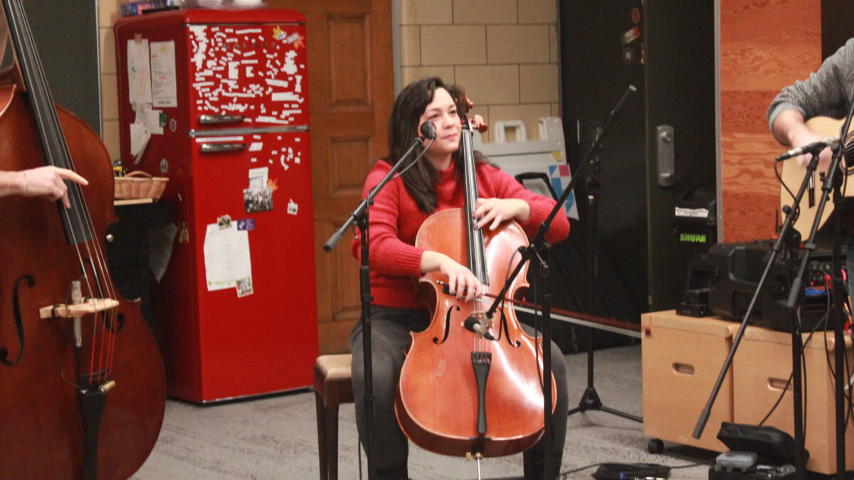 Ari-and-the-Buffalo-Kings-1 An adult playing a cello while sitting behind a microphone and in front of a red refrigerator