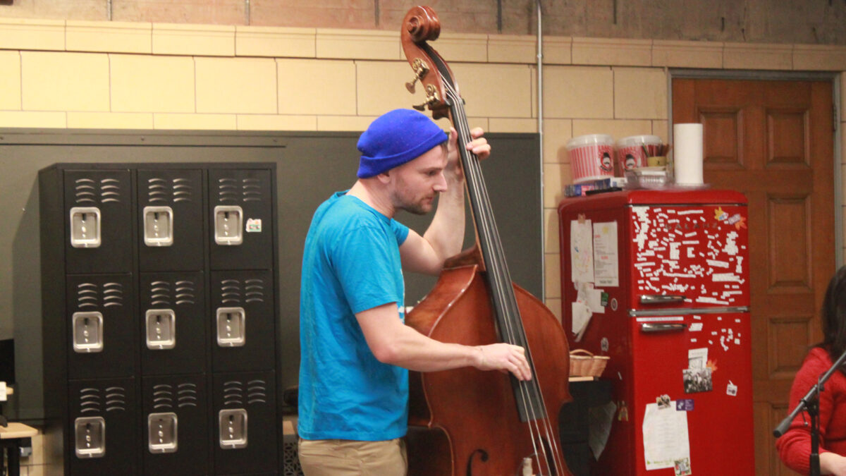 Ari-and-the-Buffalo-Kings-2 An adult playing an upright bass standing in front of black lockers and a red refrigerator