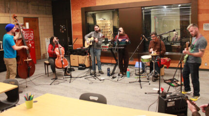 An adult playing upright bass, an adult playing cello, an adult playing acoustic guitar, an adult singing into a microphone, an adult playing a small drum set, and an adult playing electric guitar