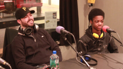 An adult and a youth wearing headphones around their necks speaking into microphones in a radio studio