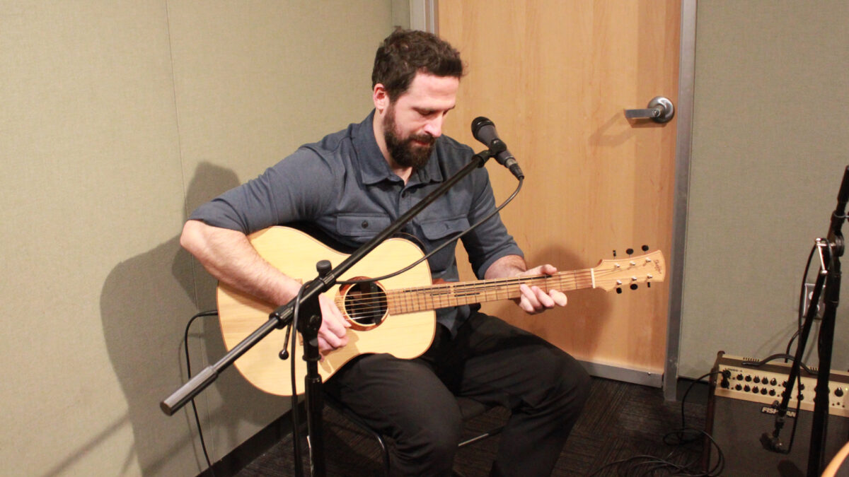 An adult playing an acoustic guitar sitting behind a microphone in a radio studio