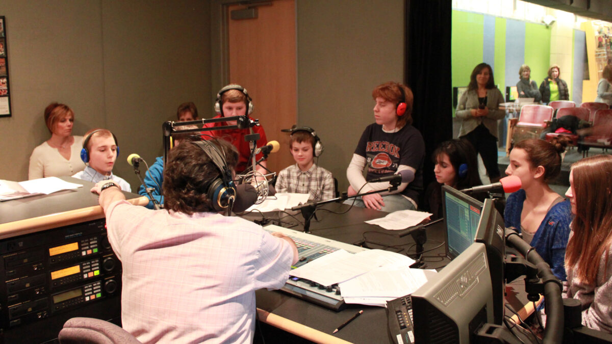 Eight youth wearing headphones speaking into microphones being interviewed by an adult wearing headphones sitting behind a microphone and a mixer as two adults in the background listen in a radio studio
