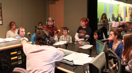 Eight youth wearing headphones speaking into microphones being interviewed by an adult wearing headphones sitting behind a microphone and a mixer as two adults in the background listen in a radio studio