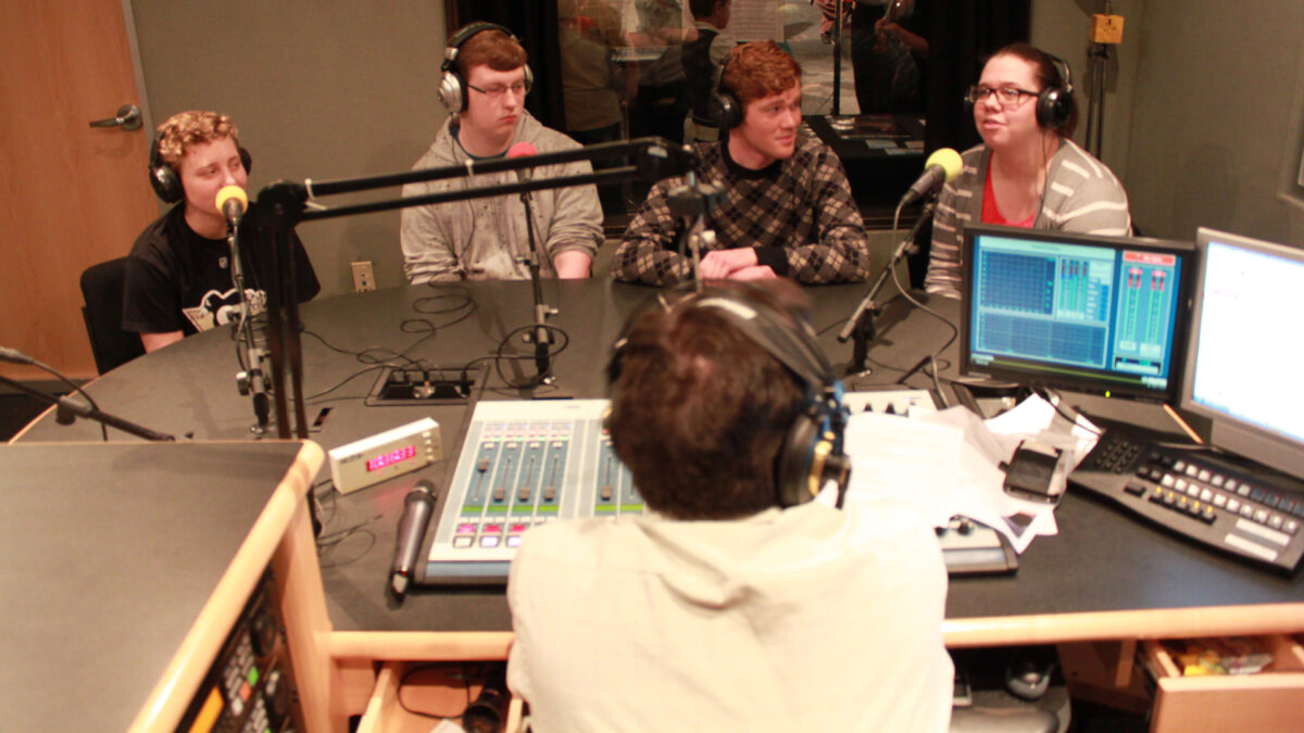 Three teens and an adult wearing headphones speaking into microphones being interviewed by an adult wearing headphones sitting behind a microphone and a mixer in a radio studio