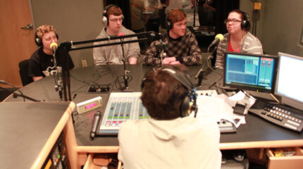 Three teens and an adult wearing headphones speaking into microphones being interviewed by an adult wearing headphones sitting behind a microphone and a mixer in a radio studio