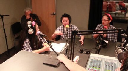 Three teens wearing headphones speaking into microphones and an adult holding a camera taking pictures in a radio studio