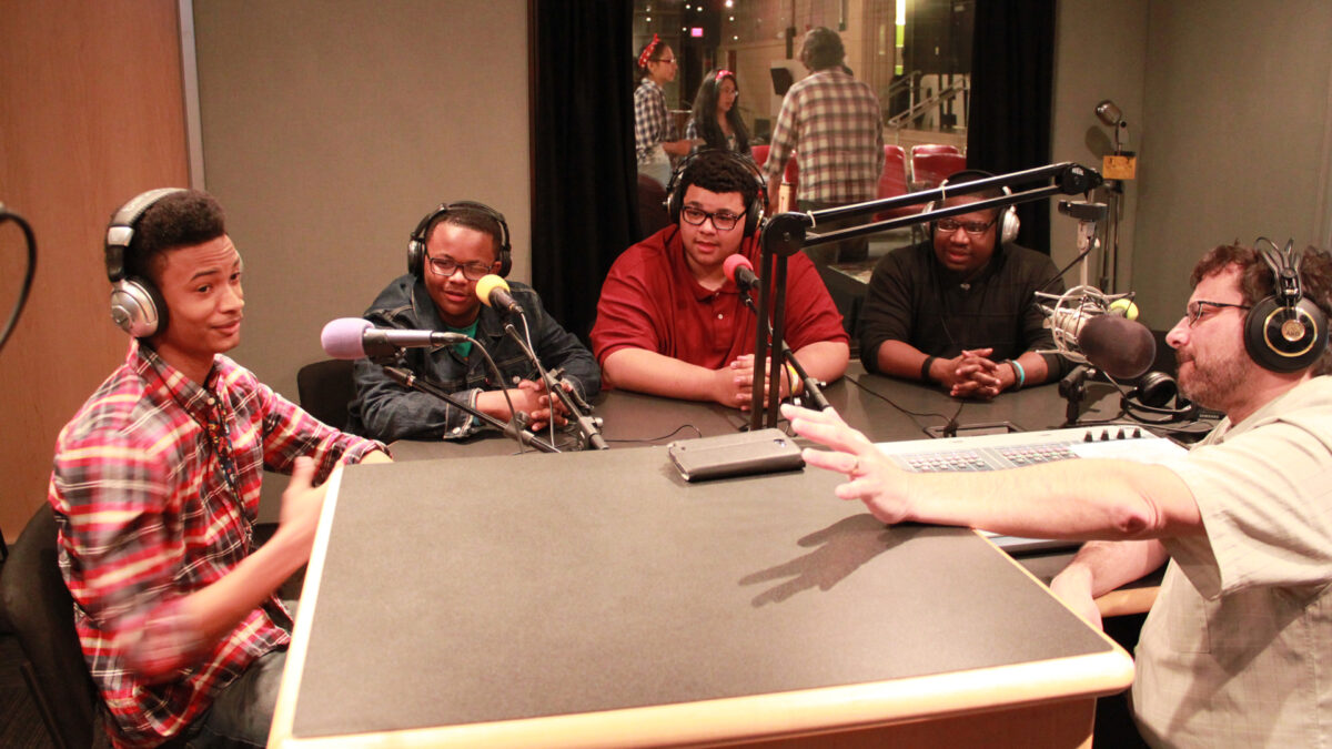 Three teens and an adult wearing headphones speaking into microphones being interviewed by an adult wearing headphones and sitting behind a microphone and a mixer in a radio studio