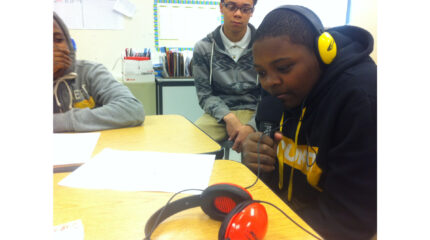 Two youth looking at another youth wearing headphones and holding a microphone while sitting at a classroom desk
