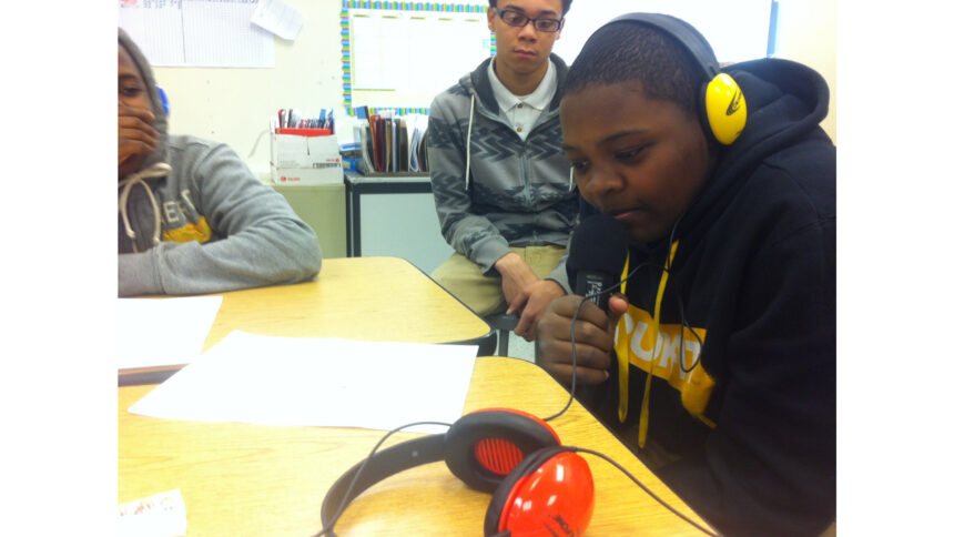 Two youth looking at another youth wearing headphones and holding a microphone while sitting at a classroom desk