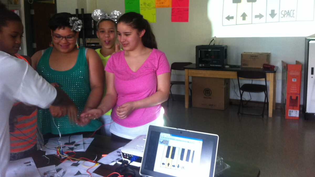 Four youths looking down at wires on a classroom desk, one of the youths sticking out their tongue at the camera