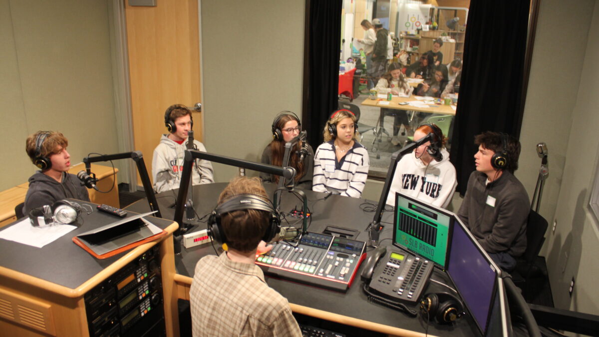 Six teens wearing headphones speaking into microphones and an adult wearing headphones sitting behind a microphone and a mixer in a radio studio
