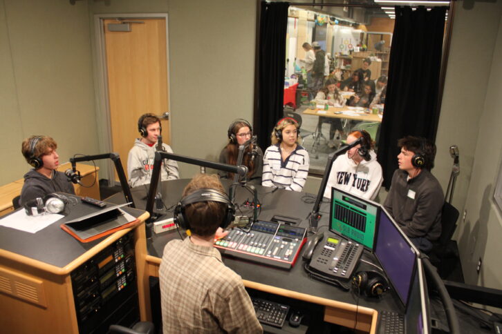 Six teens wearing headphones speaking into microphones and an adult wearing headphones sitting behind a microphone and a mixer in a radio studio
