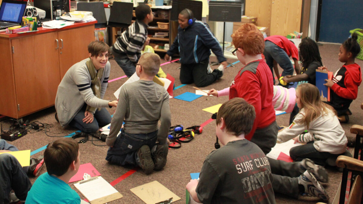 Ten youth looking at an adult, all sitting on the ground in a circle around headphones