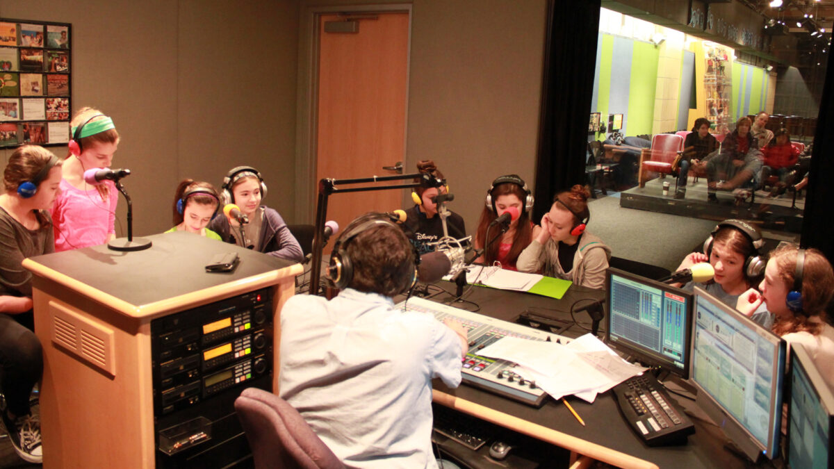 Nine youth wearing headphones speaking into microphones being interviewed by an adult wearing headphones and sitting behind a microphone and a mixing board in a radio studio