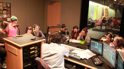 Nine youth wearing headphones speaking into microphones being interviewed by an adult wearing headphones and sitting behind a microphone and a mixing board in a radio studio