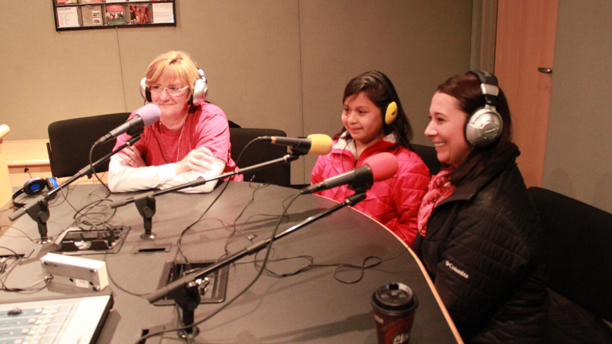 Two adults and a youth wearing headphones speaking into microphones in a radio studio