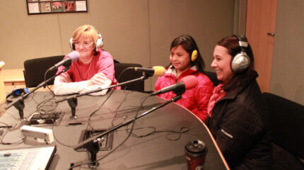 Two adults and a youth wearing headphones speaking into microphones in a radio studio