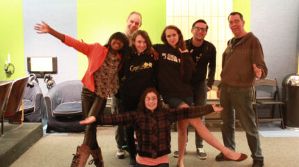 Four teens and three adults smiling and posing in profile in front of sofas and a green wall