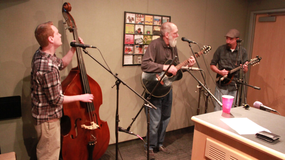 An adult playing upright bass standing behind a microphone, an adult playing acoustic guitar and singing into a microphone, and an adult playing mandolin and standing behind a microphone in a radio studio