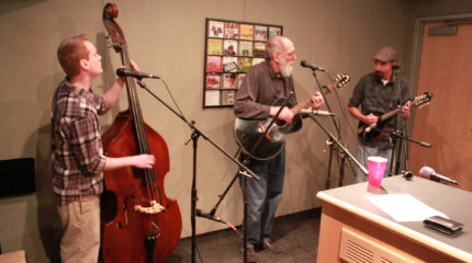 An adult playing upright bass standing behind a microphone, an adult playing acoustic guitar and singing into a microphone, and an adult playing mandolin and standing behind a microphone in a radio studio