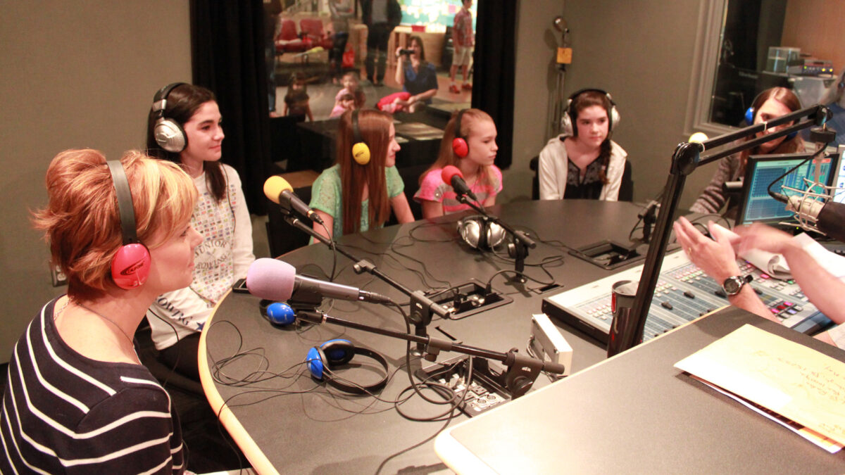 Six youth wearing headphones sitting behind microphones being interviewed in a radio studio