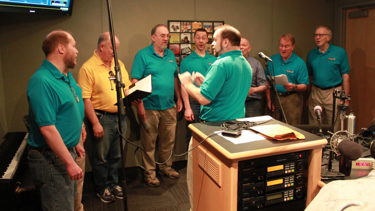 Seven adults singing in a choir and an adult conducting them in a radio studio