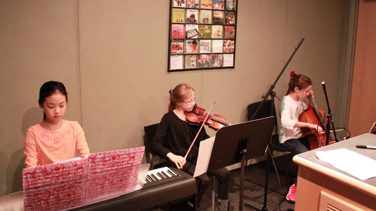 A youth playing a piano keyboard, a youth playing violin, and a youth playing cello, all looking at sheet music in a radio studio
