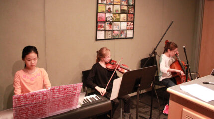 A youth playing a piano keyboard, a youth playing violin, and a youth playing cello, all looking at sheet music in a radio studio