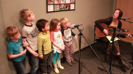 Five youth standing behind a microphone and an adult playing an acoustic guitar and singing into a microphone in a radio studio