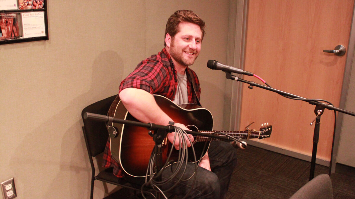An adult playing an acoustic guitar and singing into a microphone in a radio studio