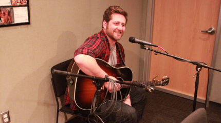 An adult playing an acoustic guitar and singing into a microphone in a radio studio