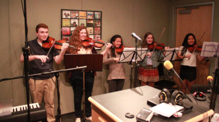 Five teens smiling while holding violins in playing position standing behind sheet music on music stands in a radio studio
