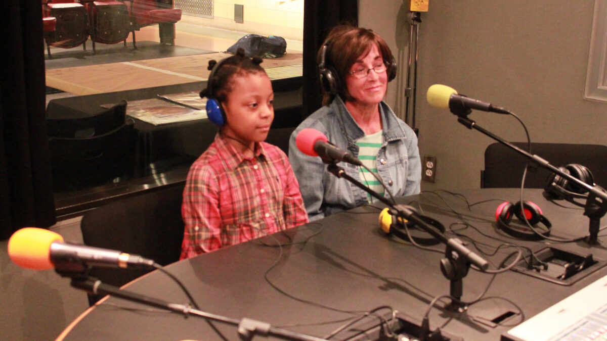 An adult and a youth wearing headphones sitting behind microphones in a radio studio