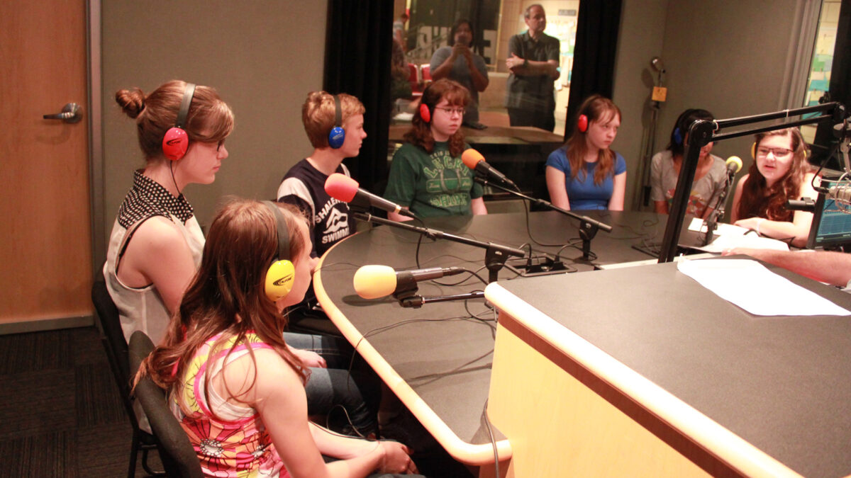 Seven youth wearing headphones speaking into microphones in a radio studio