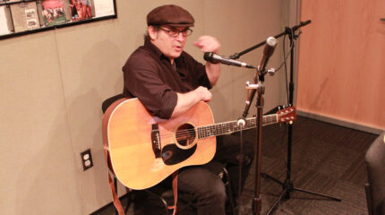 An adult holding an acoustic guitar and speaking into a microphone while gesturing with their hand in a radio studio