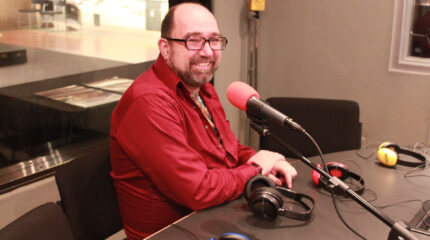 An adult smiling while sitting behind a microphone in a radio studio