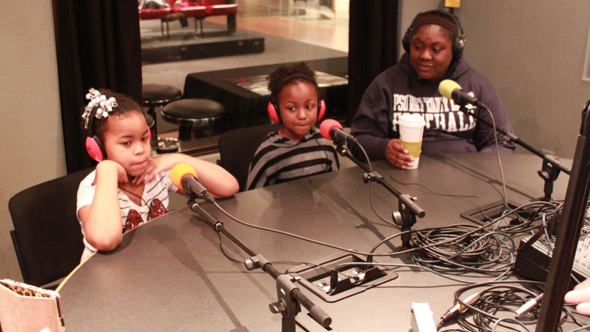 An adult and two youth wearing headphones sitting behind microphones in a radio studio