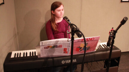 A youth sitting behind a piano keyboard, sheet music, and a microphone in a radio studio