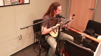 An adult playing ukulele and sitting behind a microphone in a radio studio