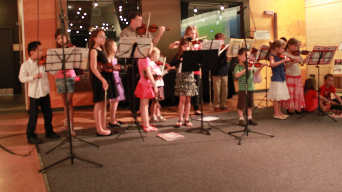 Sixteen youth and two adults with violins behind music stands in front of a radio studio