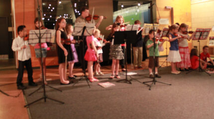 Sixteen youth and two adults with violins behind music stands in front of a radio studio