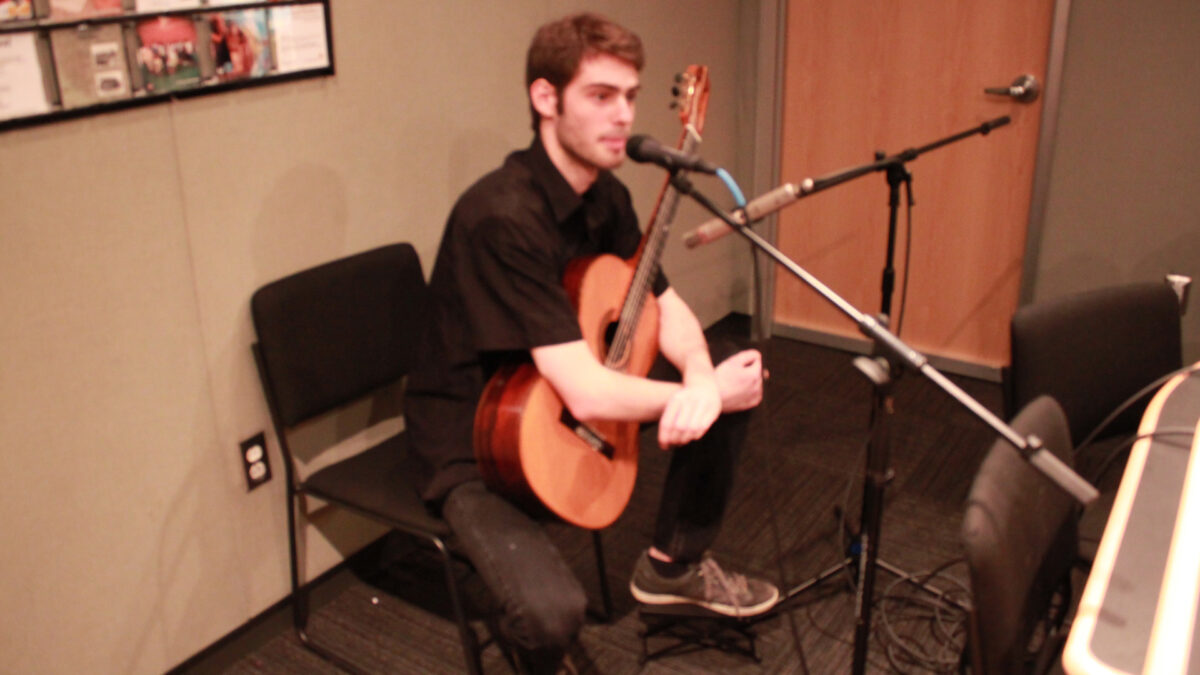 A teen holding an acoustic guitar sitting behind a microphone in a radio studio